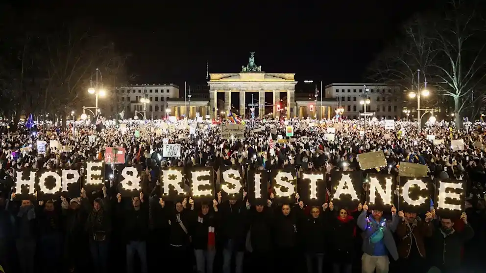 A large nighttime protest crowd in front of the Brandenburg Gate holds up illuminated signs spelling out 'HOPE &…