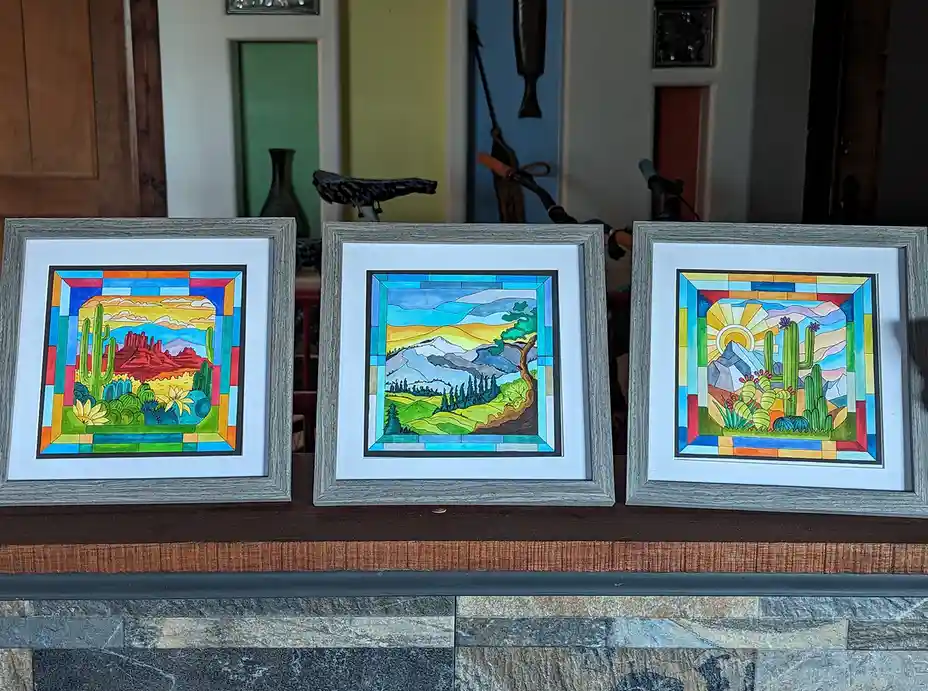 Three framed, colorful, stylized landscape paintings resembling stained glass are displayed side-by-side on a wooden mantel…