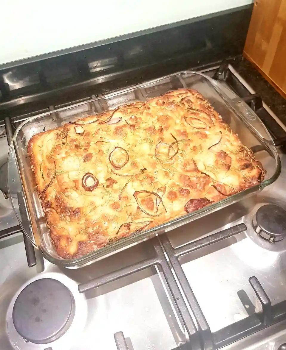A golden-brown baked dish in a glass casserole dish sits on a stovetop, with caramelized onion slices arranged to resemble a…