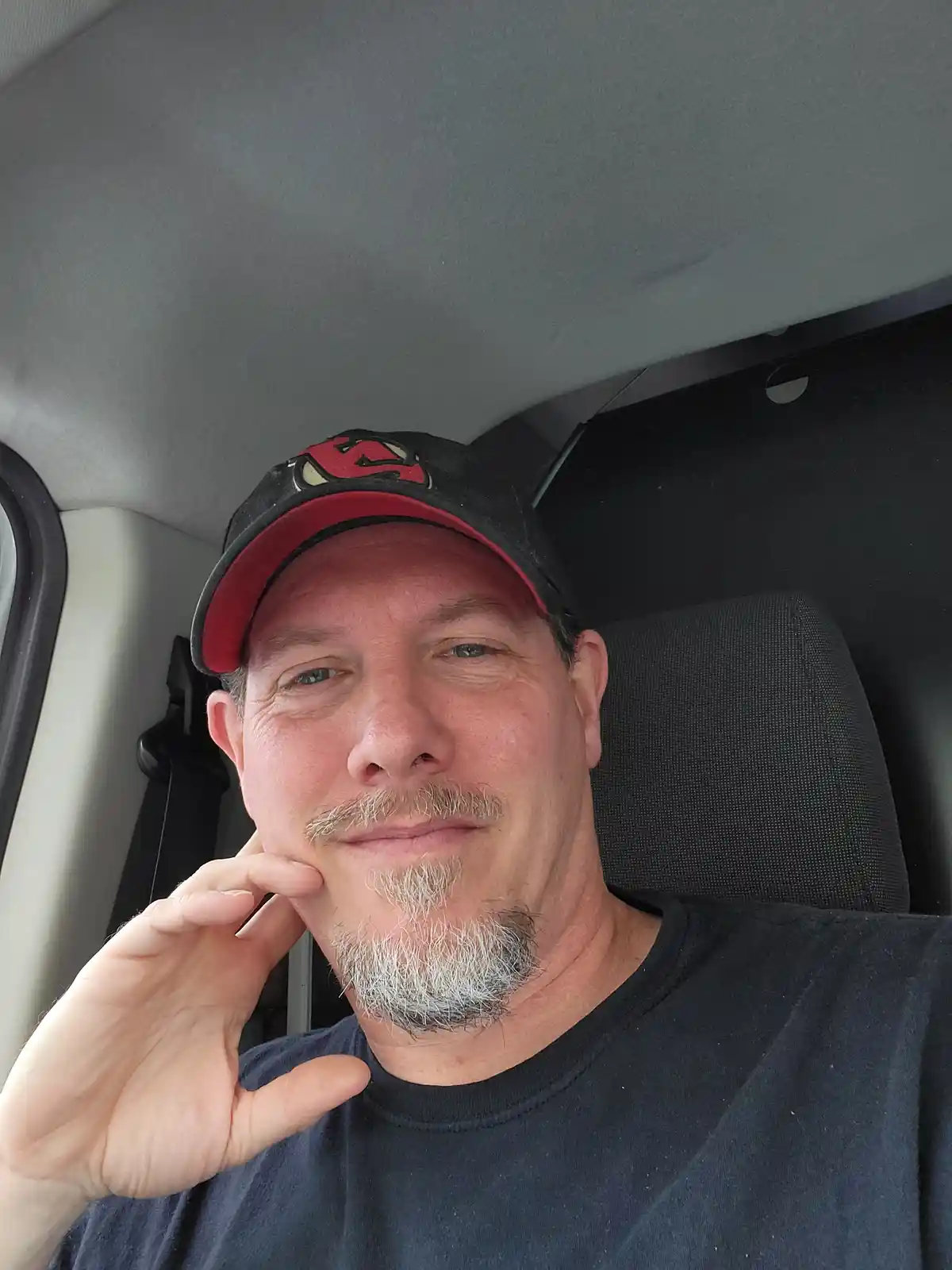 A middle-aged man with a graying goatee and mustache sits in a car, wearing a black and red baseball cap and a dark t-shirt…