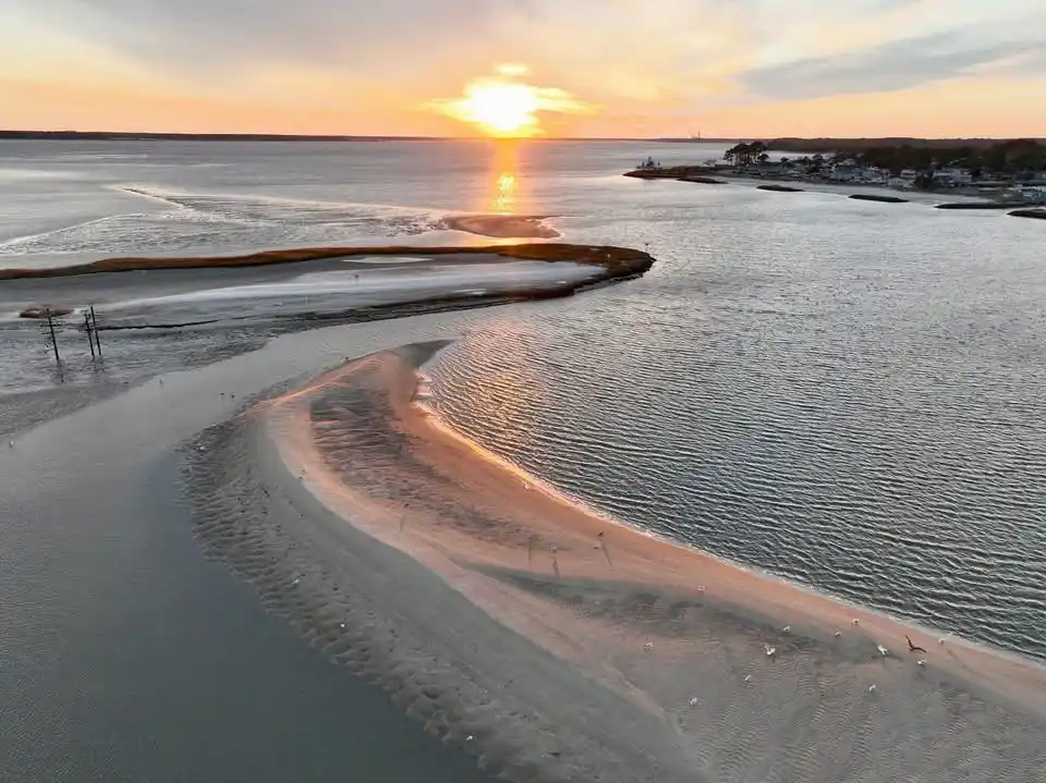 An aerial photo of a winding sandbar at sunset, with the sun low on the horizon casting golden light across the water and…