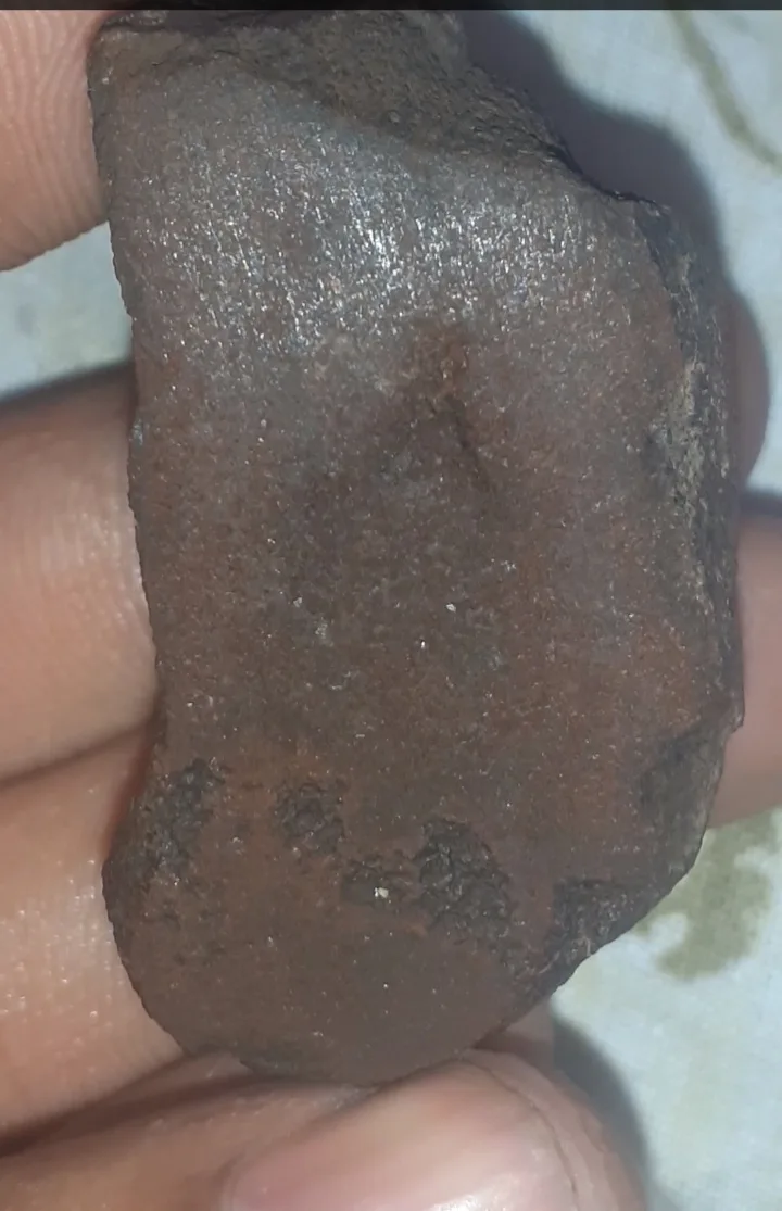 a close-up photo shows a hand holding a rough, brownish-red, irregularly shaped rock with a slightly shiny surface, against…