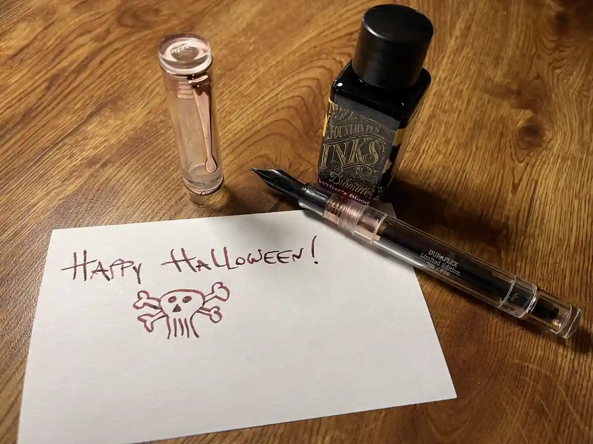 A close-up photo shows a fountain pen, a bottle of 'Writer's Blood' ink, and an empty ink converter on a wooden surface. A…