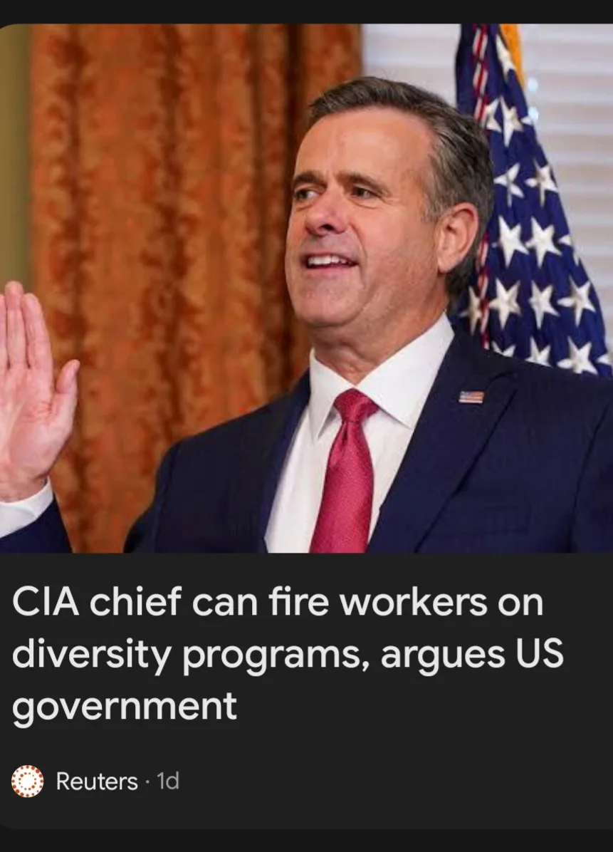A man in a dark suit and red tie raises his right hand in an oath-taking gesture, standing before an American flag and…