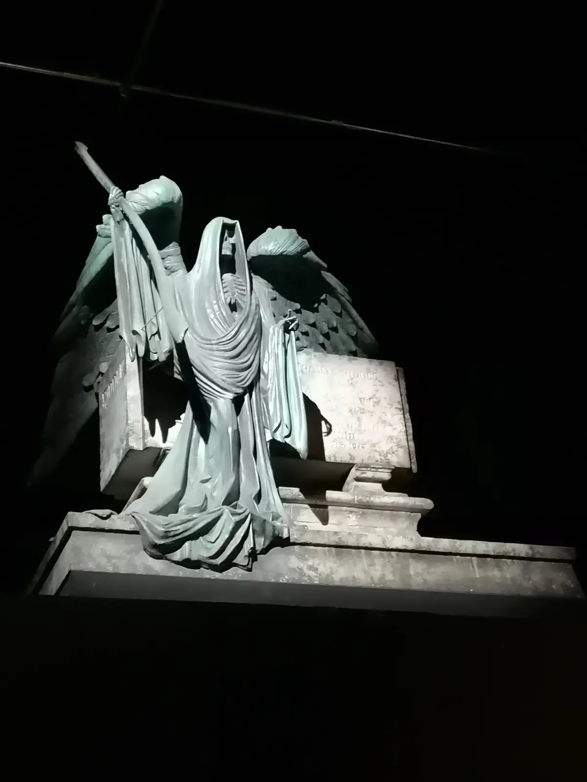 A dark, dramatic photograph shows a large, winged angel statue with a skeletal face, draped in robes, holding a staff…