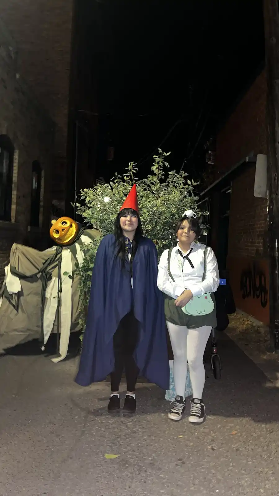 Two young women pose for a photo at night in an alleyway. The woman on the left wears a blue cape and a red pointed hat…