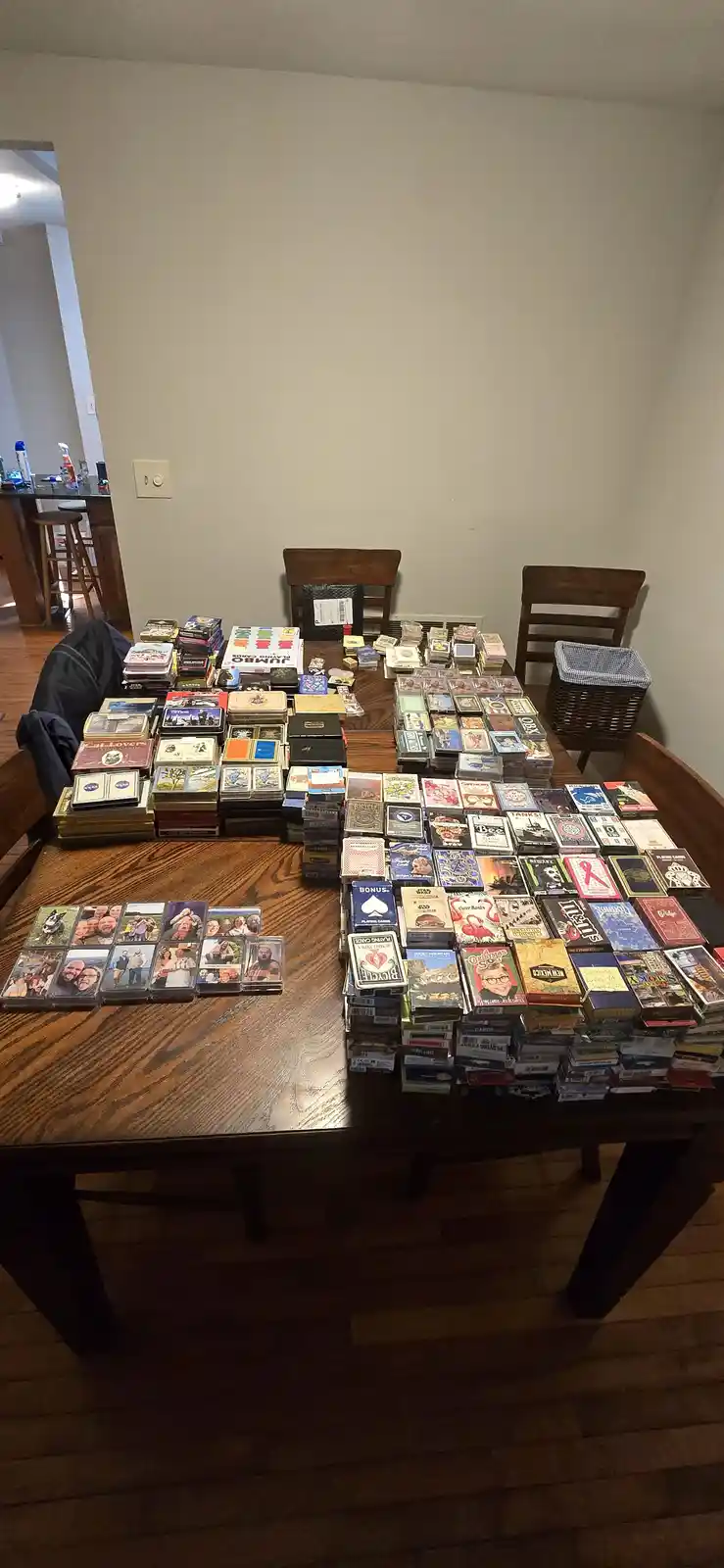 A wooden dining table is completely covered with stacks and piles of various playing card decks, including themed sets like…