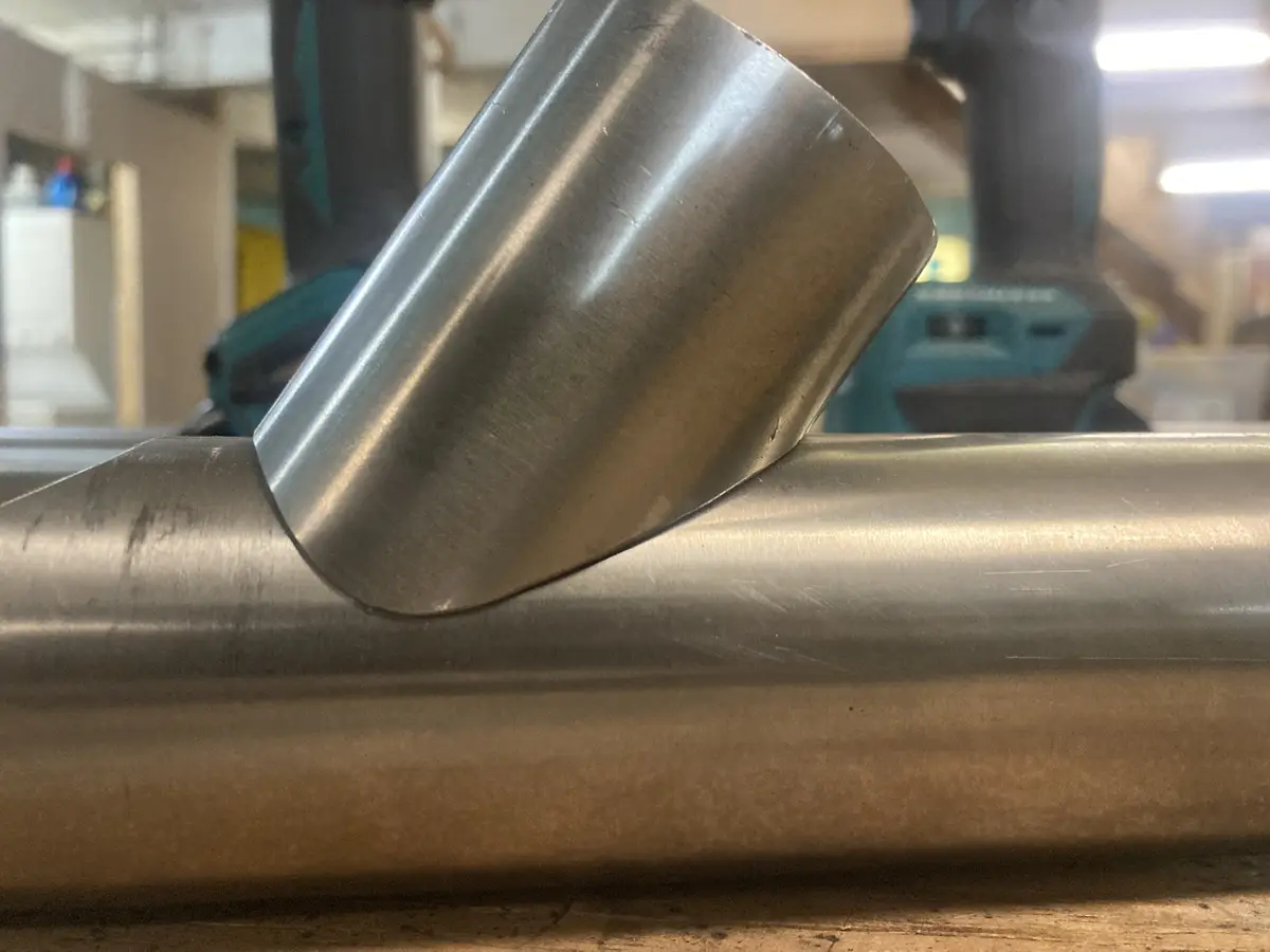 A close-up photo shows a shiny metal pipe fitting resting on a horizontal metal pipe in a workshop. In the blurred…