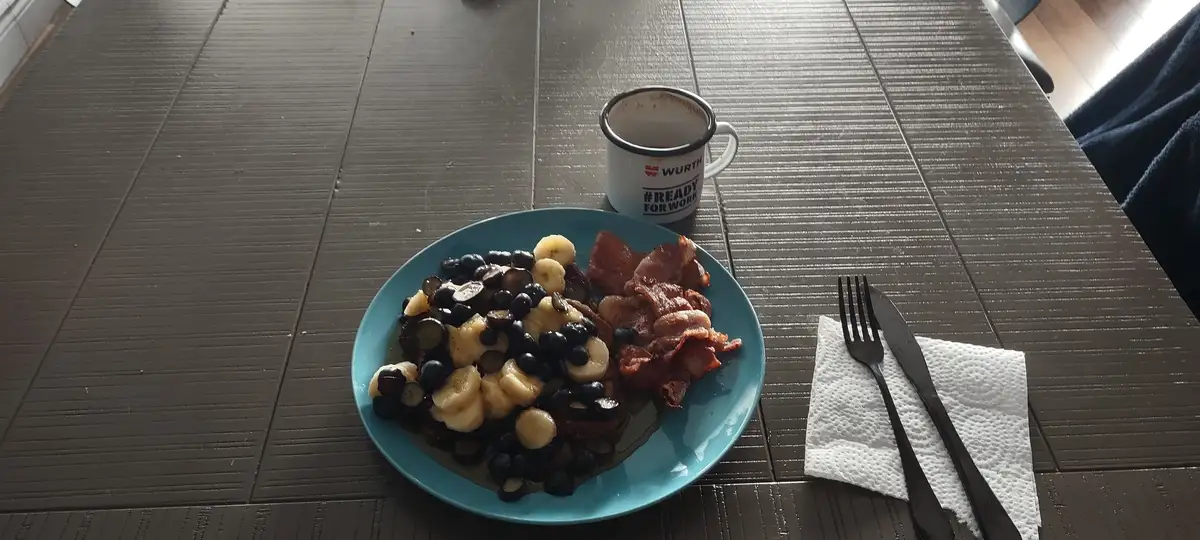 A breakfast scene shows a blue plate with pancakes topped with sliced bananas and blueberries, alongside crispy bacon. Next…