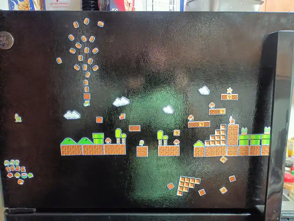 A black refrigerator door is decorated with pixelated Super Mario Bros. themed magnets, including coins, bricks, question…