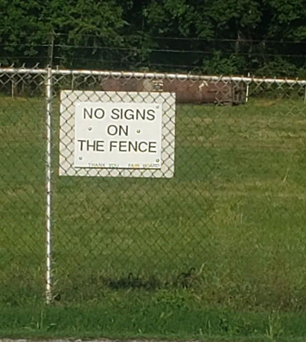 A white rectangular sign is mounted on a chain-link fence in a grassy field, displaying the text 'NO SIGNS ON THE FENCE' in…
