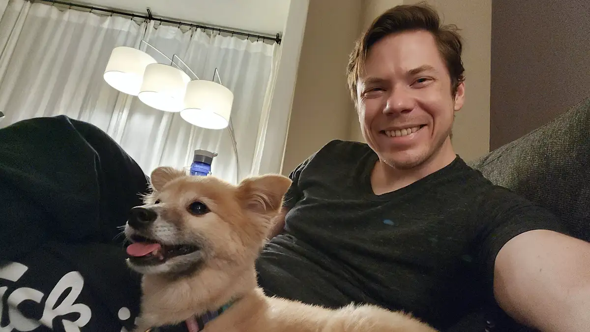 A man in a dark t-shirt smiles at the camera while sitting on a couch with a small, fluffy, tan dog beside him. The dog has…