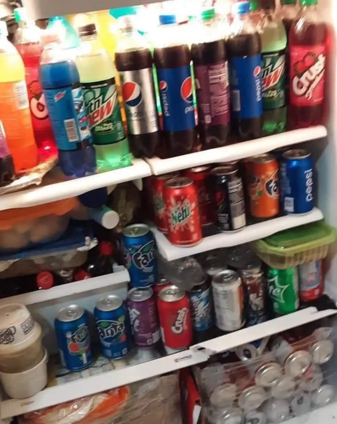A close-up photo shows a very full refrigerator with multiple shelves packed with various soda bottles and cans, including…