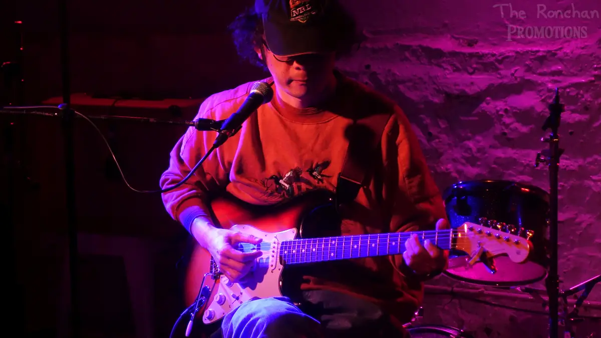 A musician with curly hair, wearing a cap and glasses, plays an electric guitar on stage under purple and red lighting. A…