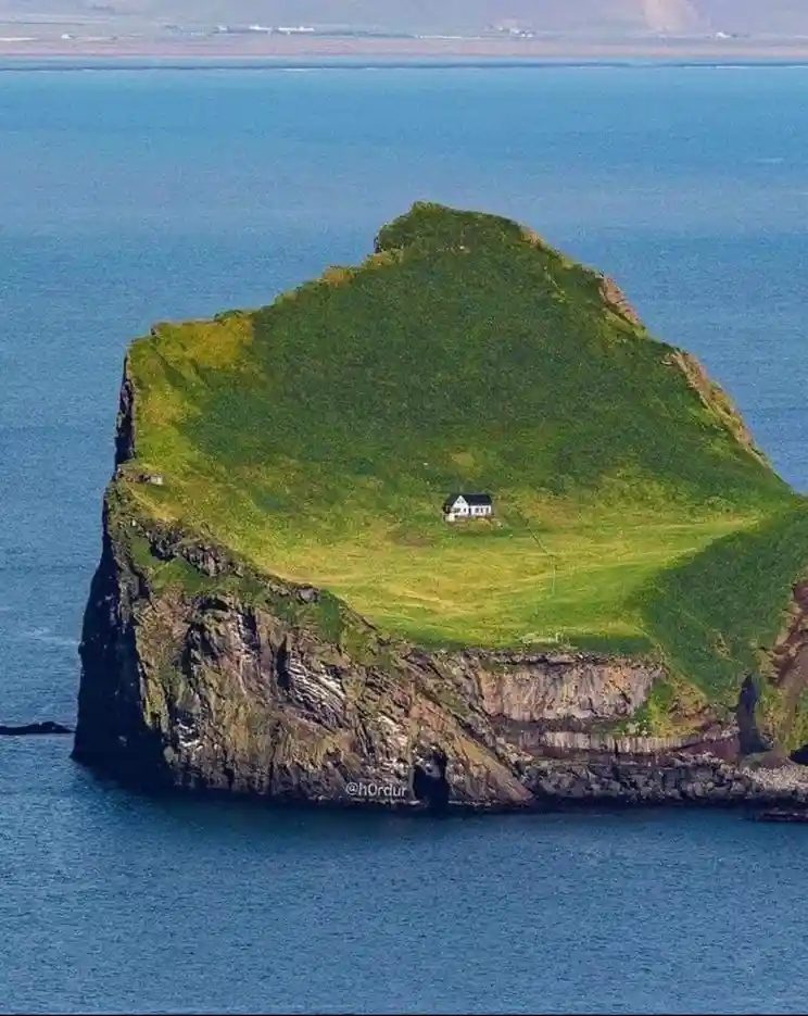 An aerial photo shows a small white house situated on a lush green, isolated cliff island surrounded by blue ocean water…