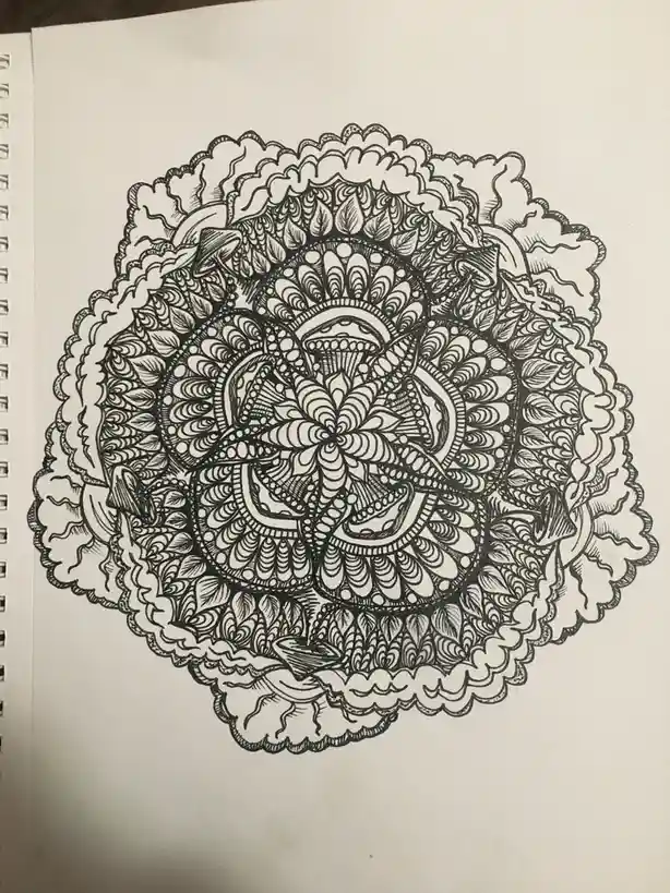 A black-and-white, highly detailed mandala drawing on a spiral-bound notebook page, featuring intricate floral and geometric…