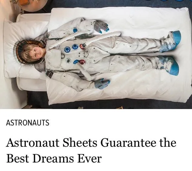 A child lies in bed wearing a full astronaut suit, including helmet and boots, while covered by white sheets. The overlaid…