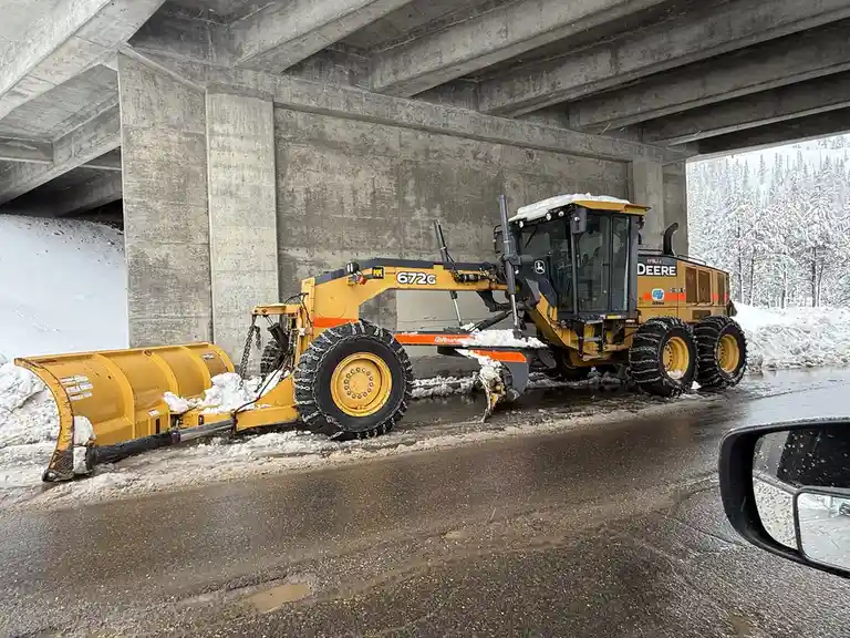 A yellow John Deere snowplow, model 672G, is parked under a concrete overpass with snow piled up around it. The vehicle has…