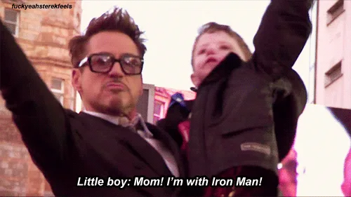 A man in a black suit and glasses holds a small child in a dark jacket while raising his arms. The child looks up excitedly…
