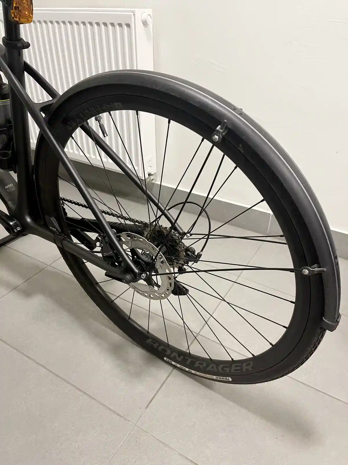 A close-up photo of a black bicycle's rear wheel, showing the tire, spokes, disc brake, and fender, parked indoors on tiled…