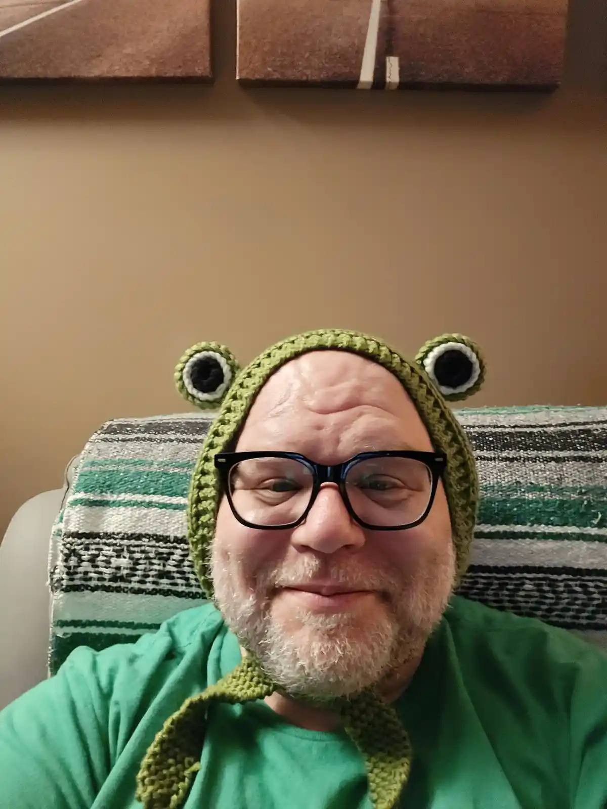 A man with a white beard and black-rimmed glasses wears a green crocheted frog hat with large googly eyes on top and a chin…