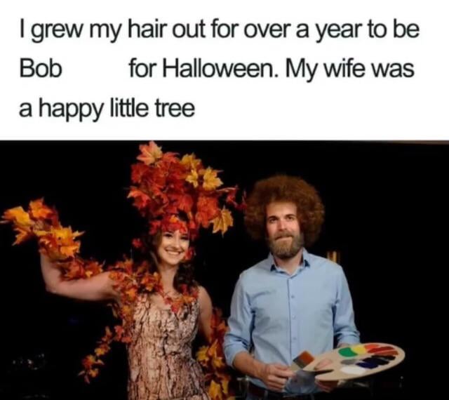 A man with a large, curly brown wig and a paint palette stands beside a woman whose hair is covered in autumn leaves and…