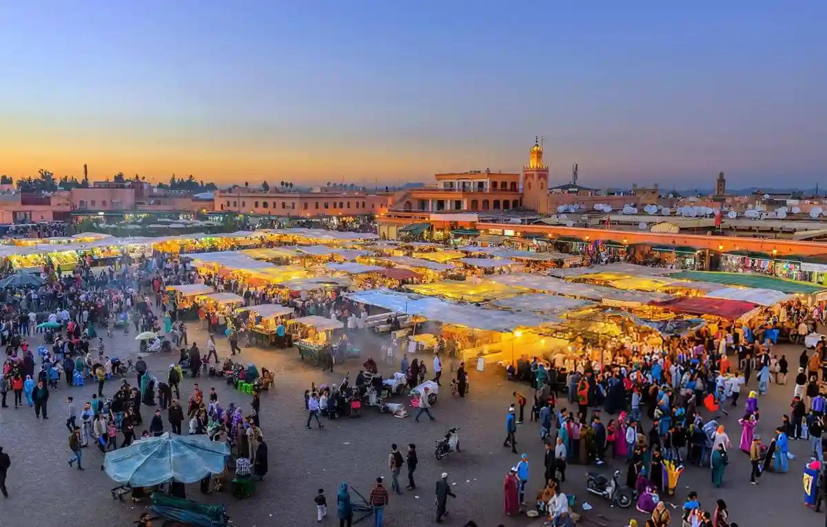 A wide-angle photo of a bustling, crowded outdoor market at dusk, with numerous stalls lit by warm lights and many people…