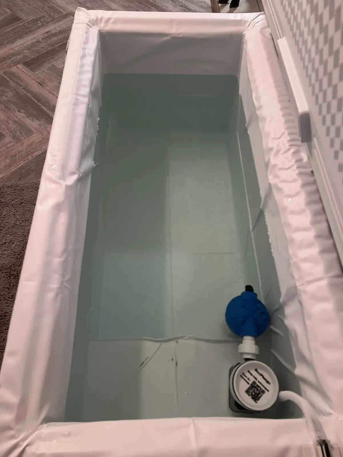 A top-down view of a bathtub filled with clear water, lined with white fabric, with a blue water heater unit and a white…