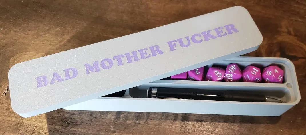 A light blue rectangular case, open on a wooden surface, contains purple dice and a black pen. The lid is engraved with the…