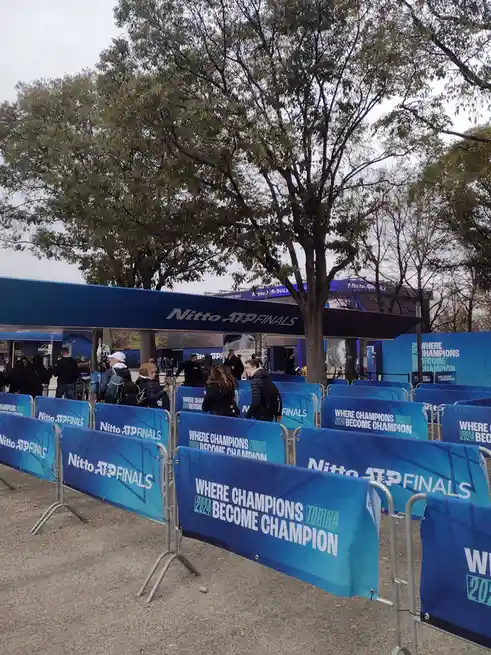 An outdoor photo shows blue event barriers and a canopy with 'Nitto ATP Finals' branding. The barriers repeatedly display…