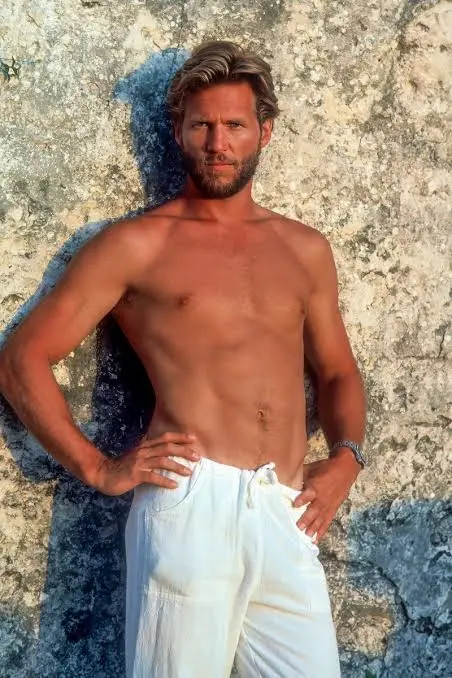 A shirtless, muscular man with a beard and styled hair stands confidently against a textured stone wall, hands on hips…