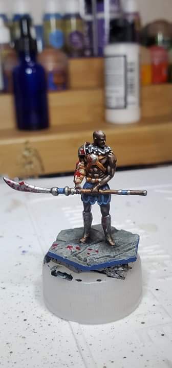A painted miniature figurine of a muscular warrior holding a large axe stands on a rocky base with red splatters resembling…