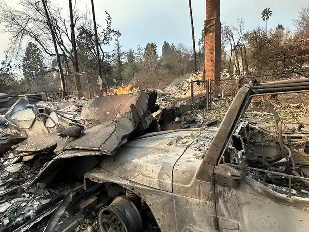 A charred, burnt-out vehicle sits amidst the rubble of a destroyed house after a wildfire, with flames still visible in the…