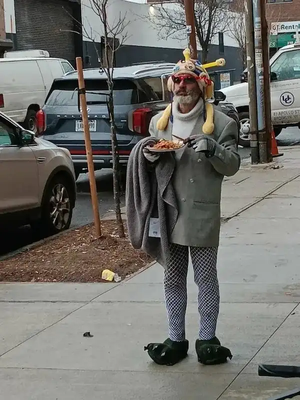 A man stands on a city sidewalk wearing a full clown costume including a colorful hat with antennae, red-rimmed glasses, a…