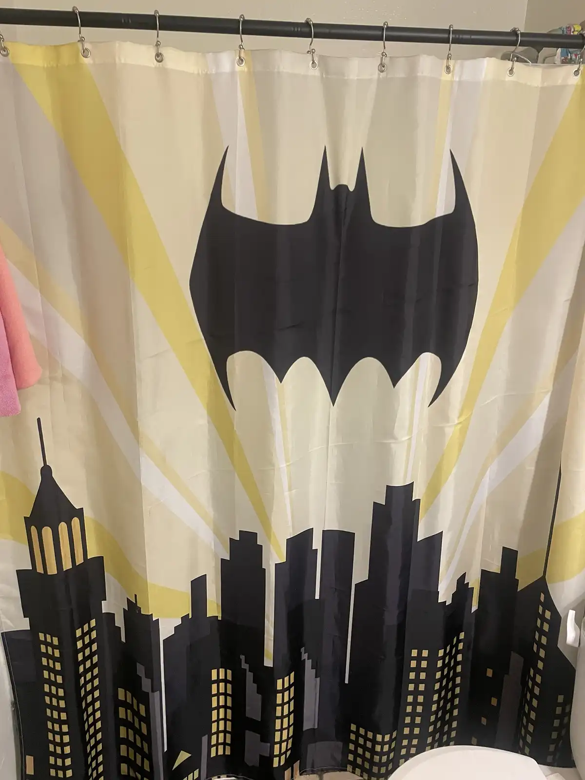 A bathroom shower curtain features a large black Batman logo centered above a stylized city skyline with illuminated…
