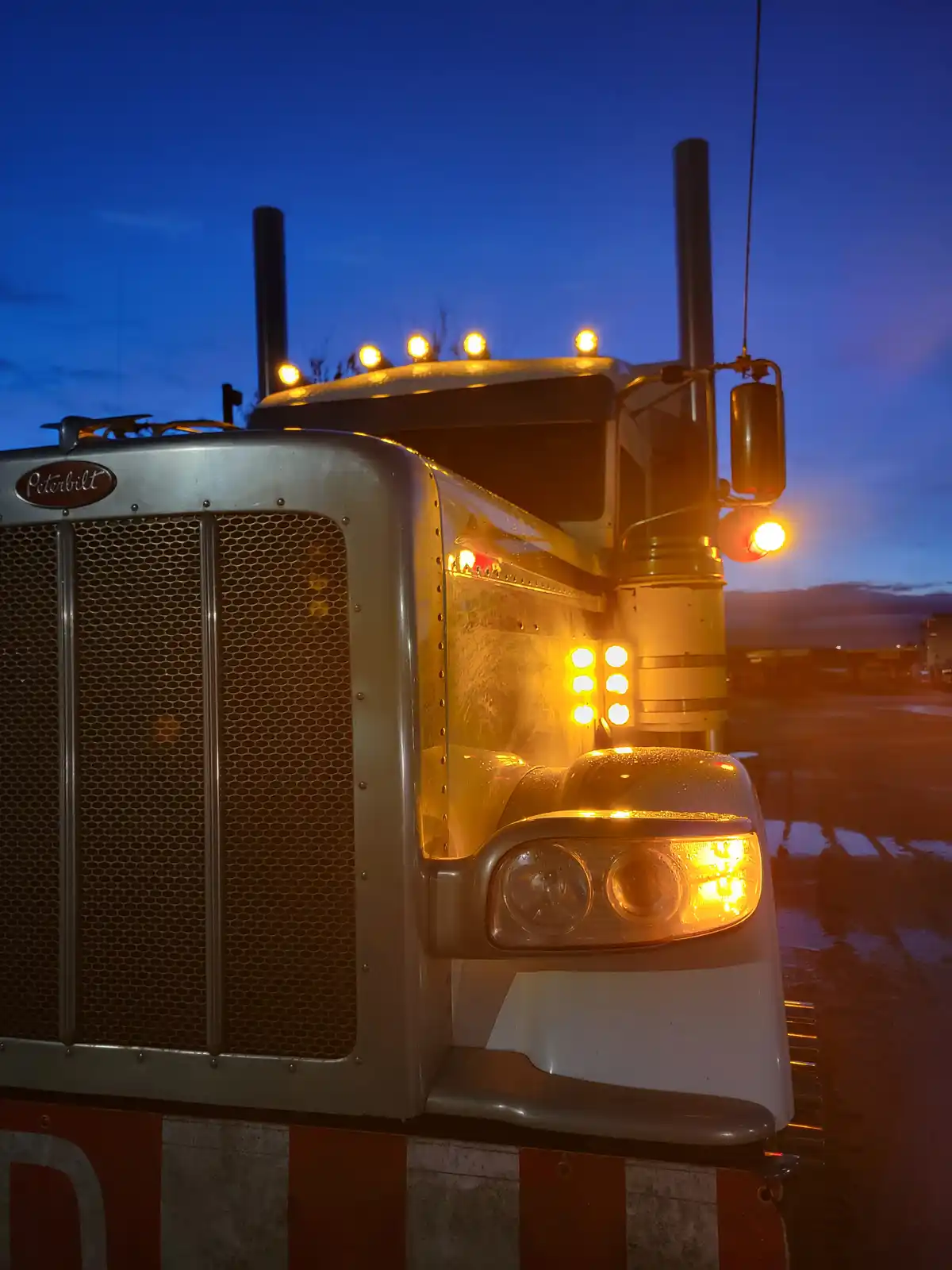 A close-up of a silver Peterbilt semi-truck's front cab at dusk, with its headlights and roof lights glowing warmly against…