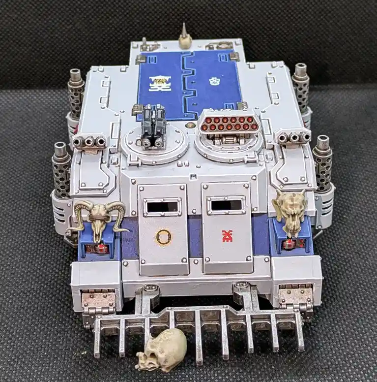 A detailed, silver and blue painted miniature tank model with skull decorations and glowing red lights, with a small skull…