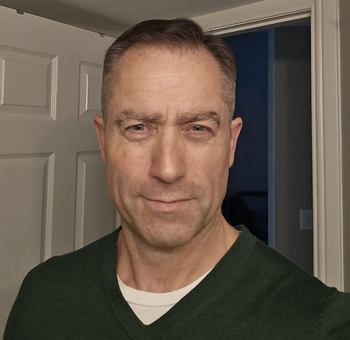 A middle-aged man with short graying hair and a stern, unamused expression stares directly at the camera. He wears a dark…