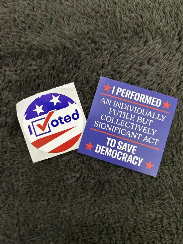Two stickers lie on a dark gray textured surface. One is the standard circular 'I Voted' sticker with stars and stripes. The…
