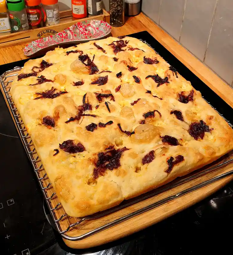 A freshly baked rectangular focaccia bread, golden-brown with puffy bubbles, sits on a wire rack atop a wooden board. Dark…
