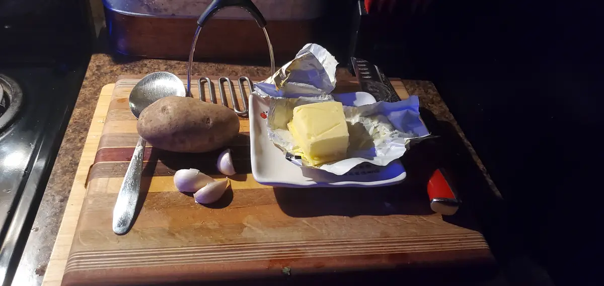 A kitchen counter shows a potato, garlic cloves, a butter pat, and cooking tools on a wooden cutting board. The overlaid…