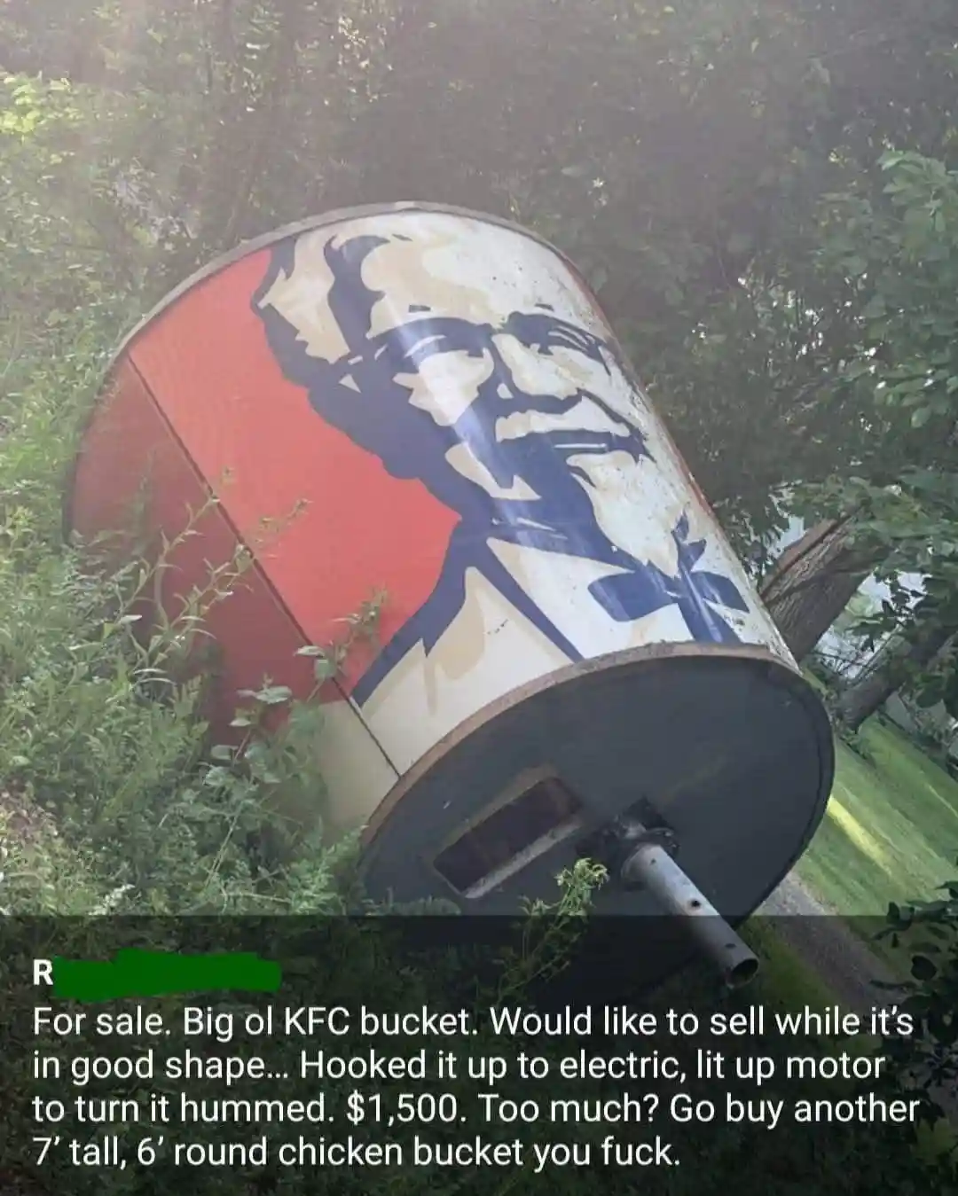 A large, weathered, cylindrical KFC bucket with Colonel Sanders' face painted on it lies on its side in overgrown grass. The…