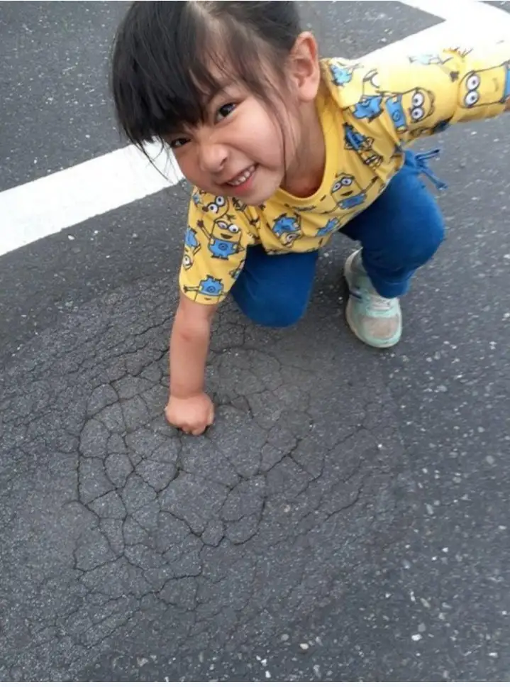 a young girl in a yellow shirt with blue minion prints and blue pants is crouching on cracked asphalt, smiling at the camera…