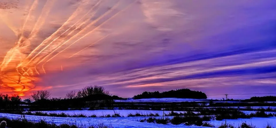 A dramatic landscape photo shows a snowy field at sunset with vibrant orange and purple skies streaked with contrails…