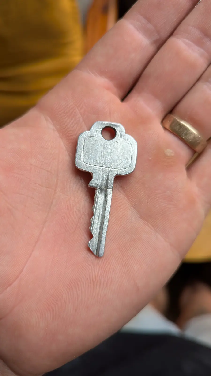 A close-up photo shows a plain metal key resting in the center of an open palm, with visible skin texture and a gold ring on…