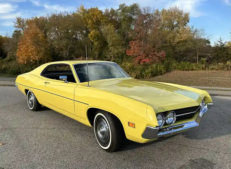 A bright yellow classic American muscle car, likely a Ford Torino, is parked on an asphalt road with autumn trees in the…