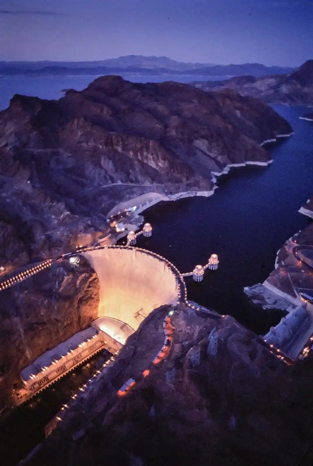 An aerial night photo shows the Hoover Dam illuminated against dark mountains and a large reservoir, with overlaid text…