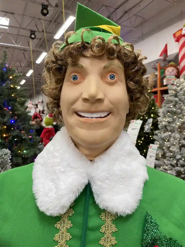 A close-up photo of a life-sized, smiling mannequin dressed as an elf from the movie 'Elf,' wearing a green hat with a…
