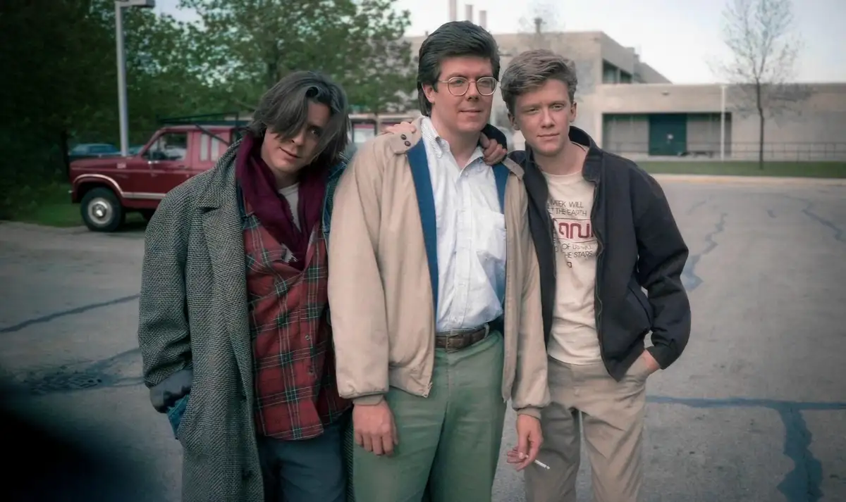 three young men in 1980s-style clothing stand together in a parking lot, with one holding a cigarette and another with a…