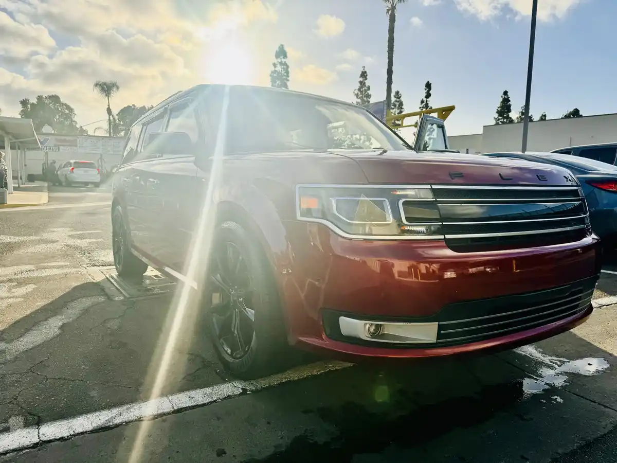 A red Ford Flex SUV is parked in a car wash lot with bright sunlight creating lens flares. The car is shiny and wet, with…